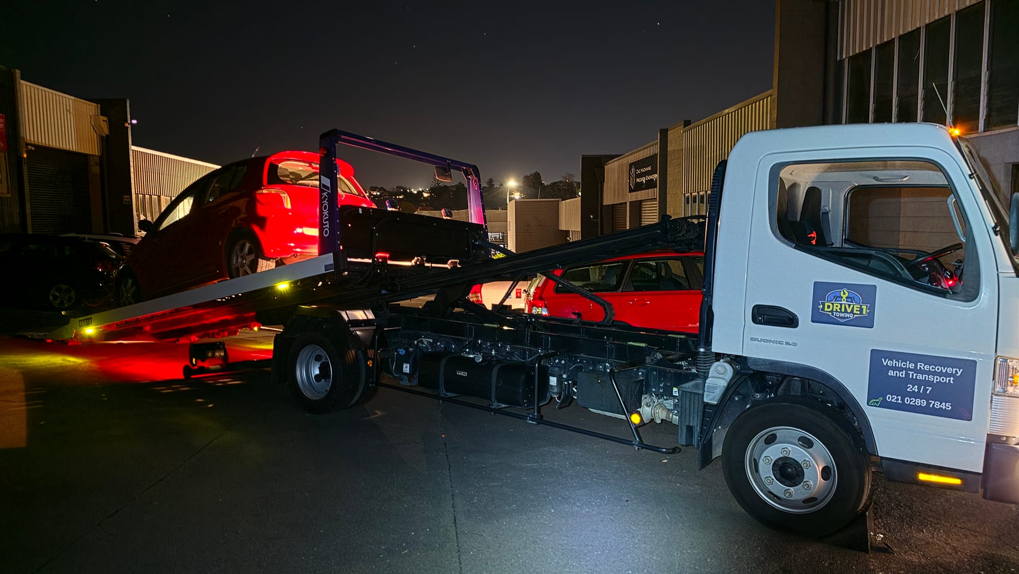 Mazda hatchback secured on a flatbed tow truck outside an Auckland dealership at night.